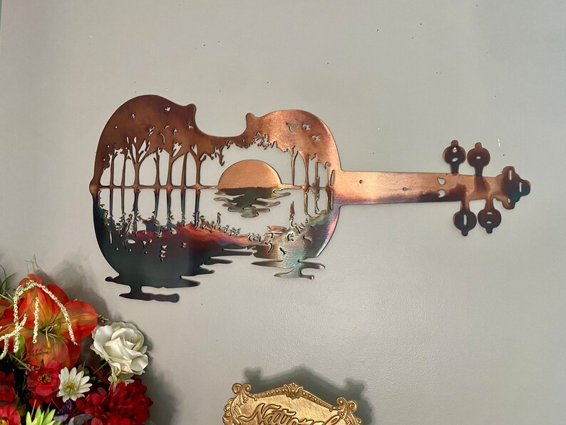 May include: A metal wall art sculpture of a guitar with a sunset scene cut out of the body. The guitar is a copper color and the scene is a silhouette of trees and a lake with a setting sun.