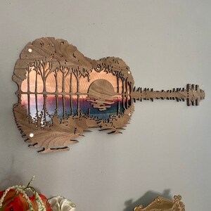 May include: A wooden guitar cutout with a sunset scene inside. The guitar is painted in shades of brown and the sunset scene is painted in shades of pink, orange, and purple.
