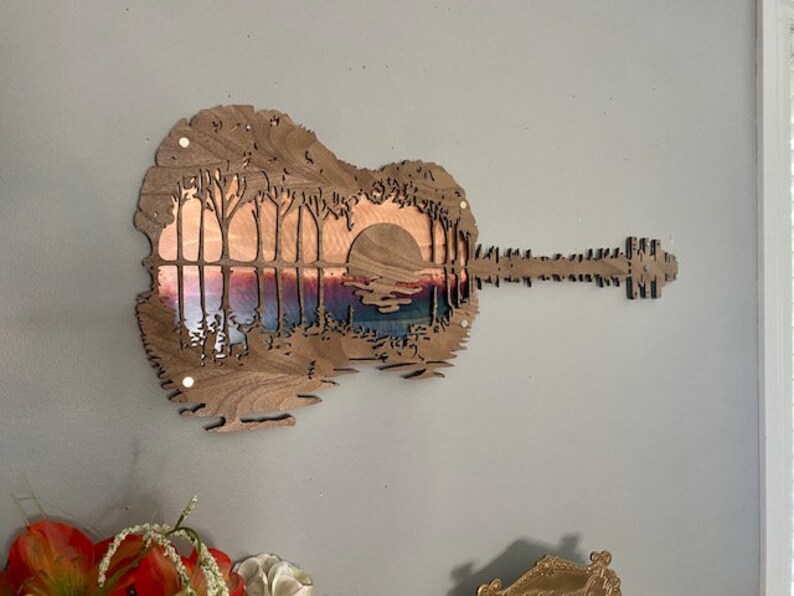 May include: A wooden guitar cutout with a sunset scene inside. The guitar is painted in shades of brown and the sunset scene is painted in shades of pink, orange, and purple.