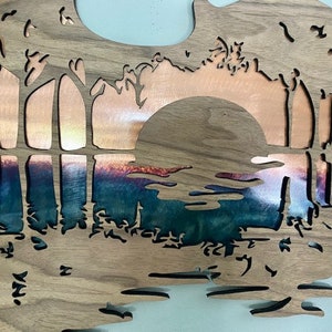 May include: A wooden wall art piece with a cut-out design of a sunset over a lake. The sunset is made of copper and the lake is a blue-green color.