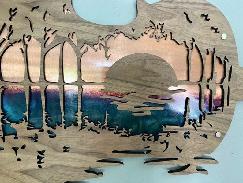 May include: A wooden wall art piece with a cut-out design of a sunset over a lake. The sunset is made of copper and the lake is a blue-green color.