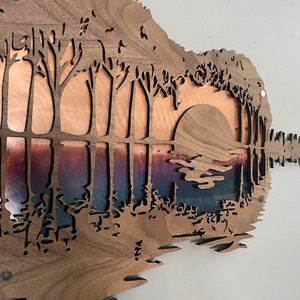 May include: A wooden guitar-shaped wall art piece with a cutout design of trees and a sunset. The background is a gradient of blue, purple, and orange.