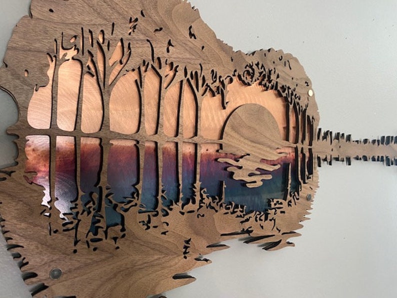 May include: A wooden guitar-shaped wall art piece with a cutout design of trees and a sunset. The background is a gradient of blue, purple, and orange.
