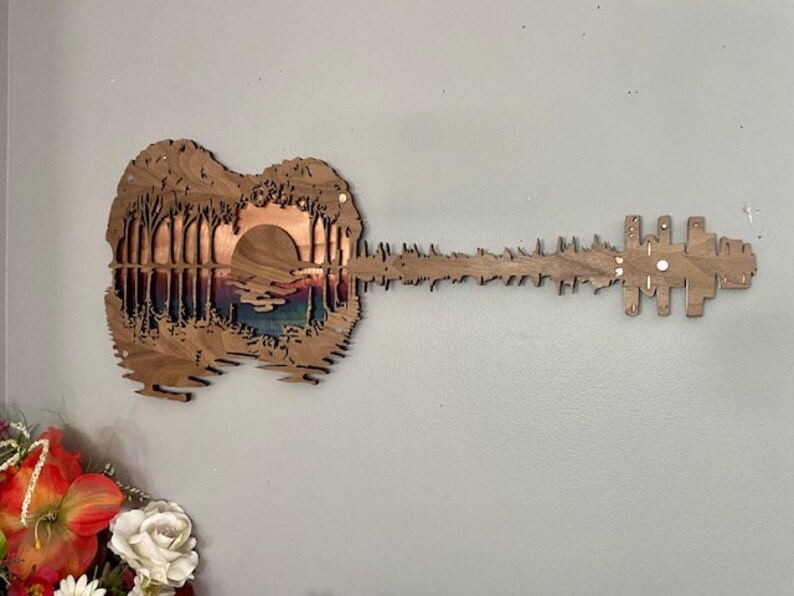 May include: A wooden wall art sculpture of a guitar with a sunset scene carved into the body. The guitar is made of dark wood and has a natural finish.