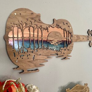 May include: A wooden wall art sculpture in the shape of a violin with a sunset scene cut out. The scene features a lake with trees on either side and a large sun in the background. The wood is a dark brown color and the scene is cut out to reveal a copper-colored background.