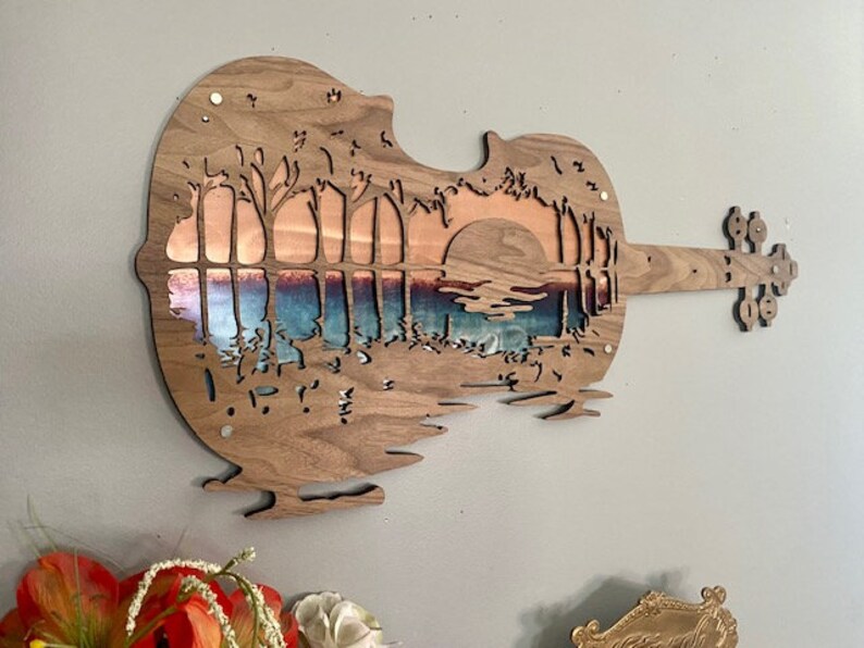 May include: A wooden wall art sculpture in the shape of a violin with a sunset scene cut out. The scene features a lake with trees on either side and a large sun in the background. The wood is a dark brown color and the scene is cut out to reveal a copper-colored background.