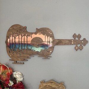 May include: A wooden violin cutout with a sunset scene carved into the body. The scene features trees, a lake, and a sun setting behind the trees. The violin is painted in shades of brown, blue, and orange.