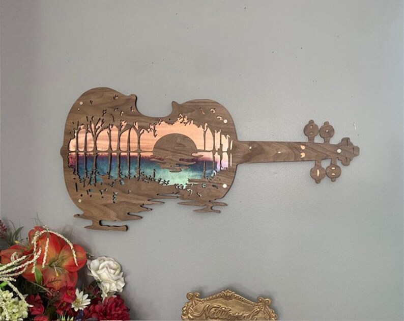 May include: A wooden violin cutout with a sunset scene carved into the body. The scene features trees, a lake, and a sun setting behind the trees. The violin is painted in shades of brown, blue, and orange.