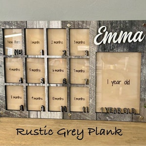 May include: Rustic grey wood photo frame with 12 openings for pictures. The frame has a white inscription that says "Emma" and "1 year old". The frame is designed to hold photos of a baby's first year.