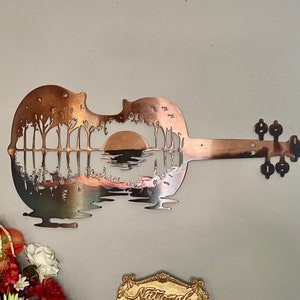 May include: A metal wall art sculpture of a guitar with a sunset and trees cut out of the body. The guitar is painted in a copper color.