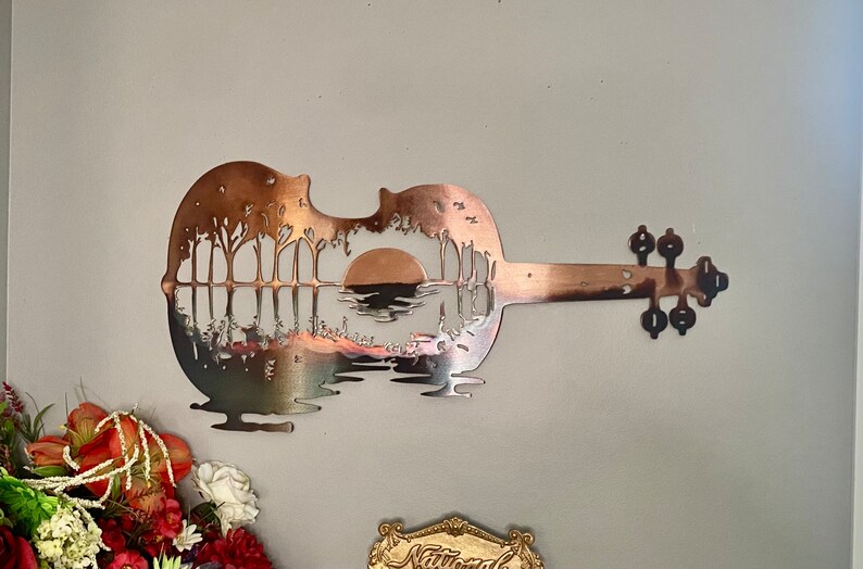 May include: A metal wall art sculpture of a guitar with a sunset and trees cut out of the body. The guitar is painted in a copper color.