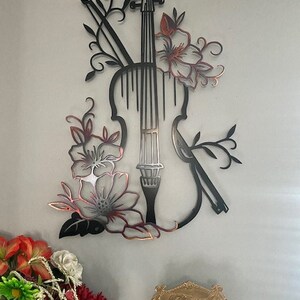 May include: Black metal wall art featuring a violin with floral accents. The violin is outlined in black and the flowers are a copper color.