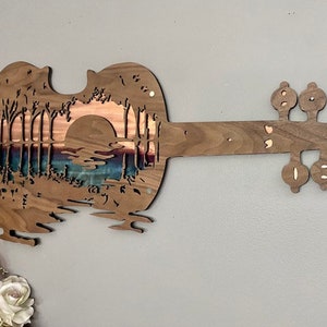 May include: A wooden wall art sculpture in the shape of a violin with a sunset scene carved into the body. The scene features a bridge, trees, and a lake.