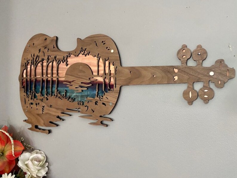 May include: A wooden wall art sculpture in the shape of a violin with a sunset scene carved into the body. The scene features a bridge, trees, and a lake.