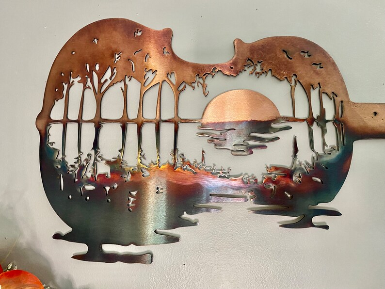 May include: A metal wall art sculpture of a sunset over a lake with trees in the foreground. The sculpture is shaped like a guitar and has a copper finish.