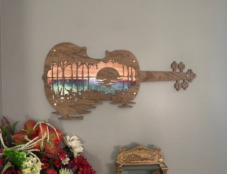 May include: A wooden cutout of a violin with a sunset scene carved into the body. The scene features a lake, trees, and a setting sun. The violin is painted with a gradient of colors, from blue to orange.