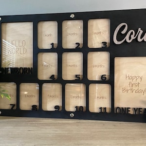 May include: Black wooden photo frame with 12 openings for pictures, each labeled with a month of the year. The frame has the words "Hello World", "Newborn", "Happy First Birthday", and "One Year" printed on it. The name "Cora" is written in silver lettering on the top right corner of the frame.