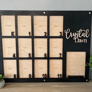 May include: Black wooden photo frame with 12 individual picture slots for each grade from kindergarten to 12th grade. The frame has the text "Crystal Dawn" and "GRADUATION" written on it.