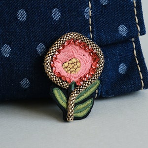 Op de afbeelding: Geborduurde bloemvormige broche met een roze bloem, gouden details en rode strass-steentjes. De bloem heeft een gouden hart en groene bladeren. De broche is geplaatst tegen een blauwe denim achtergrond met witte stippen.