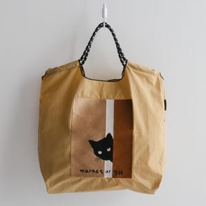 Pode incluir: Bolsa tote bege com um gráfico de gato preto espreitando por trás de um painel listrado marrom e branco. A bolsa tem alças de bolinhas pretas e brancas e o texto "market at SH".