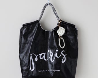 May include: Black tote bag with white script text that says "paris" and the phrase "everyday is a new beginning". The bag has a black and white patterned handle and a white flower charm with a pearl accent.
