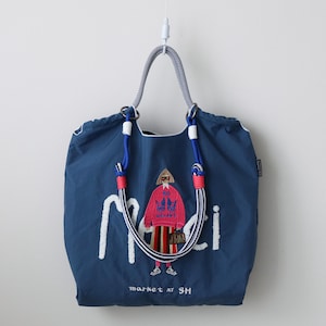 May include: A navy blue tote bag with white embroidered text and a colorful illustration of a person wearing a pink sweater. The bag has a striped handle and the text "market at SH" at the bottom.