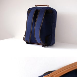 May include: A navy blue canvas backpack with brown leather accents and buckles. The backpack has a top handle and a zippered front pocket. The brand name "ROKZIM" is printed on a white label on the front of the bag.