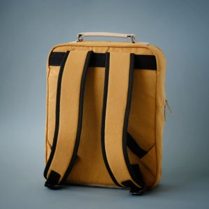 May include: A mustard yellow canvas backpack with black adjustable straps and a top handle. The backpack has a zippered pocket on the side.