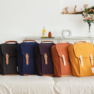 May include: Five canvas backpacks in different colors, including black, navy blue, purple, orange, and yellow. Each backpack has two adjustable straps and a buckle closure. The backpacks are lined up on a white surface.