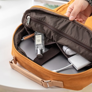 May include: A yellow canvas backpack with a brown zipper and a brown lining. The backpack is open and you can see a laptop, a pen, a bottle of perfume, and a white charger inside.