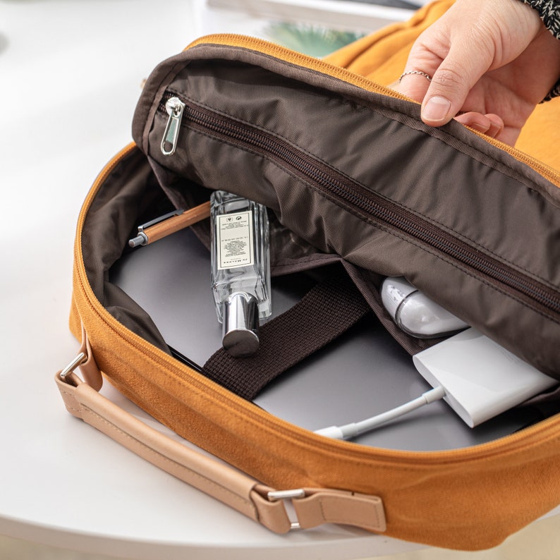 May include: A yellow canvas backpack with a brown zipper and a brown lining. The backpack is open and you can see a laptop, a pen, a bottle of perfume, and a white charger inside.