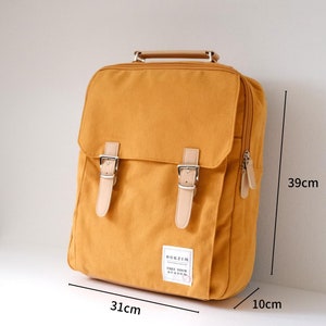 May include: A mustard yellow canvas backpack with brown leather straps and buckles. The backpack has a zipper on the side and a small patch on the front that says "Bokzim Free Your Burden". The backpack measures 39 cm tall, 31 cm wide, and 10 cm deep.