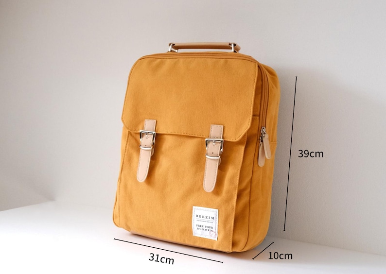 May include: A mustard yellow canvas backpack with brown leather straps and buckles. The backpack has a zipper on the side and a small patch on the front that says "Bokzim Free Your Burden". The backpack measures 39 cm tall, 31 cm wide, and 10 cm deep.