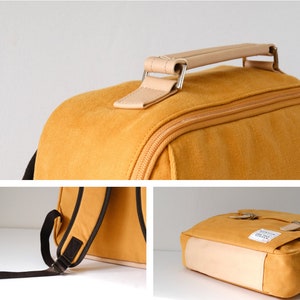 May include: A yellow canvas backpack with a brown leather handle and brown straps. The backpack has a zipper closure and a small pocket on the front.