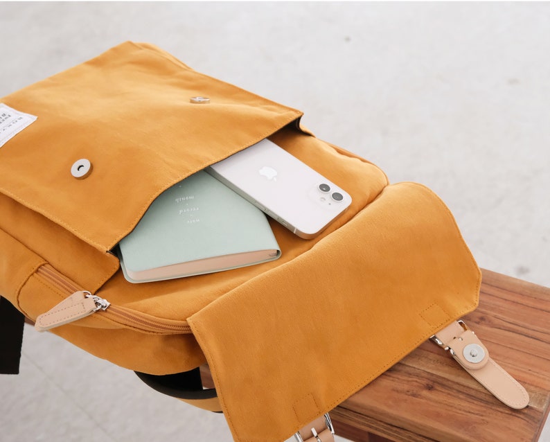 May include: A mustard yellow canvas backpack with a large flap pocket. The pocket is open and a white phone and a green notebook are inside. The backpack has a brown leather strap and a brown leather buckle.