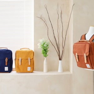 May include: Three canvas backpacks in different colors: navy blue, mustard yellow, and brown. The backpacks have two adjustable straps and two front pockets with buckles.