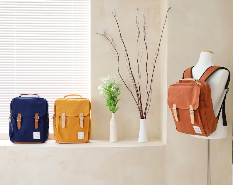 May include: Three canvas backpacks in different colors: navy blue, mustard yellow, and brown. The backpacks have two adjustable straps and two front pockets with buckles.