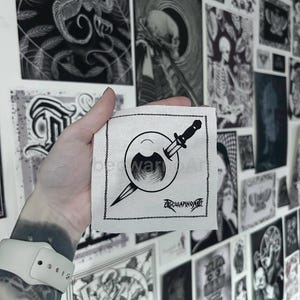 Peut inclure: Un patch carré blanc avec un graphisme noir représentant un œil percé par un poignard. Le texte "bedwar" est au-dessus de l'œil et "bedwarpaint" en dessous. Le fond présente diverses impressions d'art en noir et blanc.