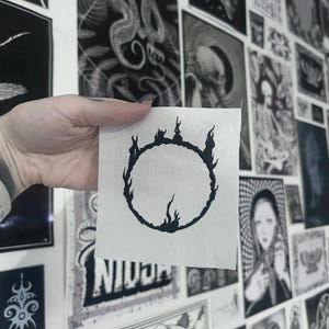 Peut inclure: Un petit patch de tissu blanc carré avec un motif circulaire noir évoquant des flammes. Le patch est présenté devant un mur orné de nombreuses impressions d'art en noir et blanc, créant un arrière-plan de style galerie.