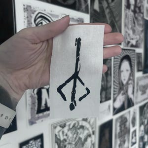 Könnte beinhalten: Ein kleines, rechteckiges Stoffstück in Weiß mit einem schwarzen, stilisierten Symbol. Das Symbol ist eine vertikale Linie mit einer kreuzartigen Form oben und einer tropfenförmigen Form unten. Das Patch wird in einer Hand gehalten, mit anderen Schwarz-Weiß-Kunstwerken im Hintergrund.