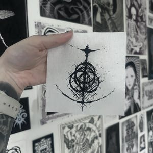 Peut inclure: Un petit patch de tissu blanc carré avec un motif abstrait noir. Le motif comprend une structure centrale en forme de toile avec des lignes rayonnantes et une figure en haut. Le patch est tenu devant un mur couvert d'estampes en noir et blanc.