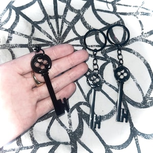 May include: Black keychains with a vintage key design. The keys have a circular top with four holes and a long, rectangular shaft. The keychains are made of metal and have a matte finish. The background is a white surface with a black spiderweb design.
