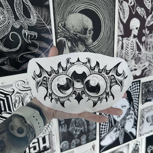 May include: A white, oval decorative stone with a black eye design. The artwork features three large eyes with detailed elements and a decorative border. The background includes various black and white art prints.