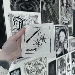 Peut inclure: Un petit carré de tissu blanc cassé avec une illustration noire d'une arme de fléau. Le fléau comporte deux boules à pointes reliées par des chaînes à une poignée. Le texte "BERWARNOART" est en dessous. Le tissu est bordé d'un cadre noir. Le fond présente diverses impressions d'art en noir et blanc.