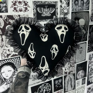 Könnte beinhalten: Ein herzförmiges schwarzes Kissen mit weißen Scream-Masken-Grafiken. Das Kissen hat eine gerüschte schwarze Bordüre. Der Hintergrund zeigt verschiedene Schwarz-Weiß-Illustrationen und Kunstwerke.