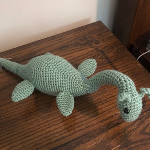 May include: A crocheted green plush toy shaped like a Loch Ness Monster. The toy has a long neck, a small head with black eyes, and four fins.