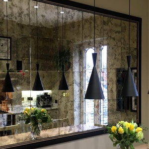 May include: A large rectangular mirror with a black frame and a distressed antique finish. The mirror is divided into multiple square panels. Four black pendant lights hang from the ceiling in front of the mirror.