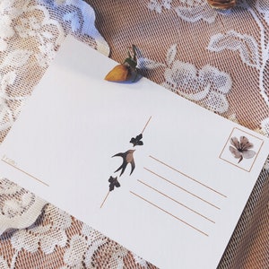 May include: A blank white postcard with a brown bird and leaves design. The postcard has a space for a stamp and an address. The postcard is lying on a white lace tablecloth.