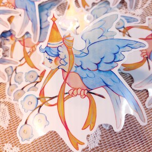 May include: A blue bird with a yellow crown and a yellow ribbon is perched on a branch with white flowers. The bird has a pink body and blue wings. The background is white.