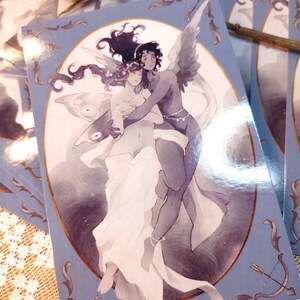 May include: A blue and gold postcard featuring a watercolor illustration of two figures embracing. The figure on the left has butterfly wings and the figure on the right has angel wings. The background is a soft blue with a gold border.